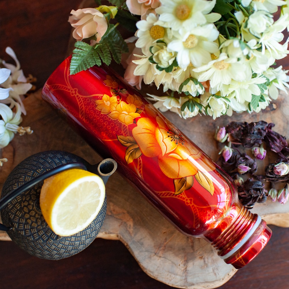Rote Kupferflasche mit Blumenmuster auf Holz umgeben von Blumen und Teepott mit Zitrone