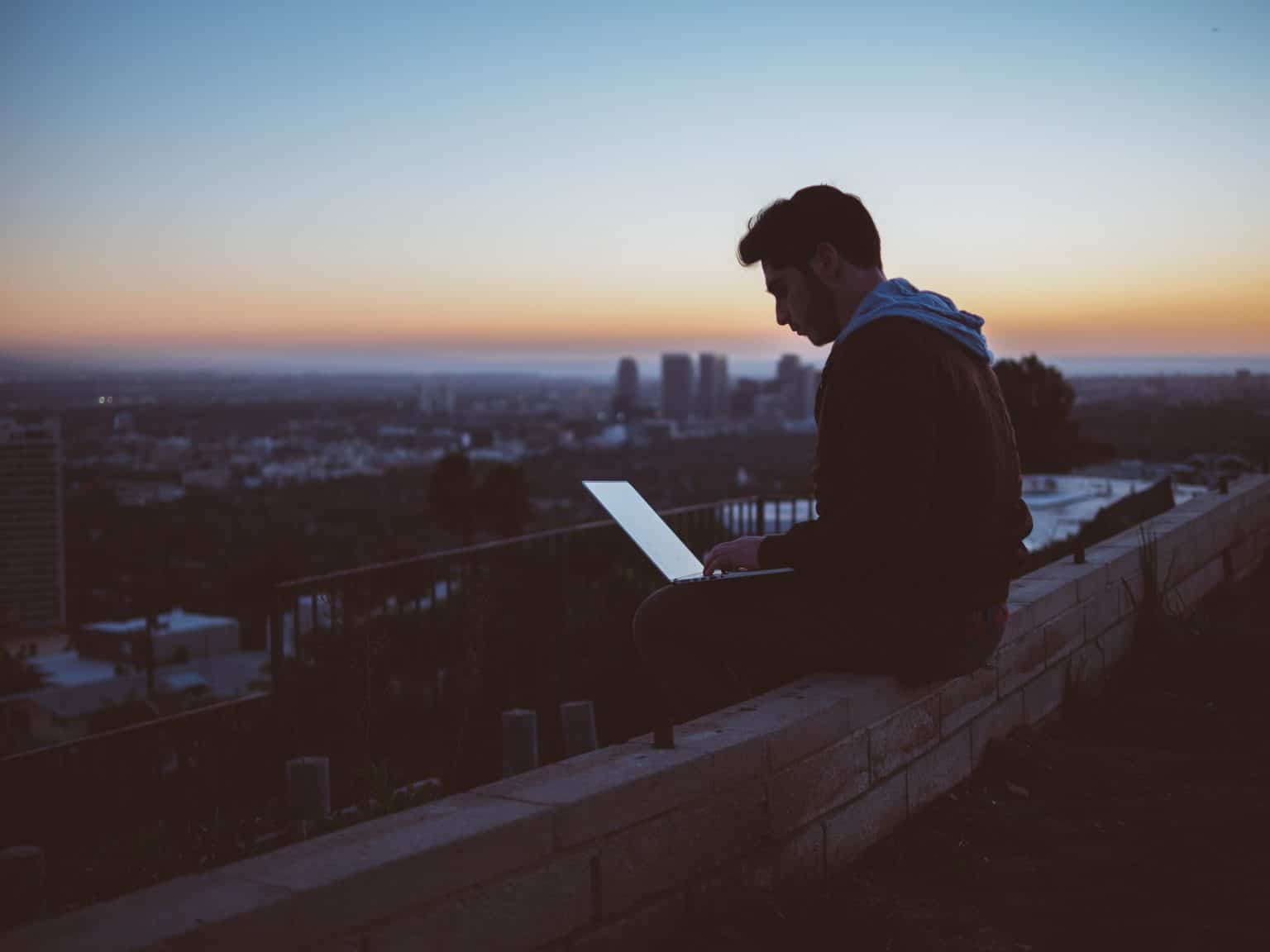 Mann sitzt auf Mauer mit Laptop vor Skyline und Sonnenuntergang