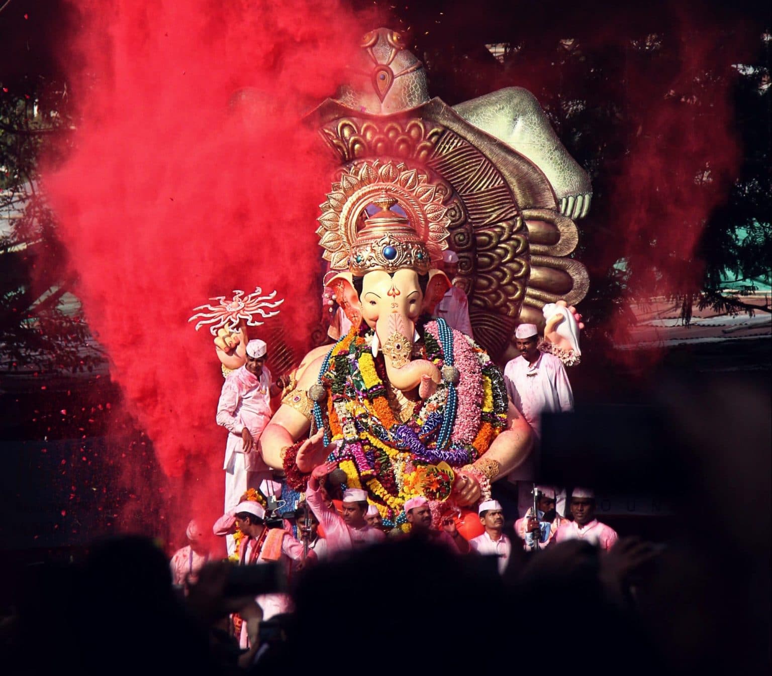 Ganesha Statue getragen von mehreren Männern in weißer Kleidung mit Blumenketten und rotem Holi Pulver