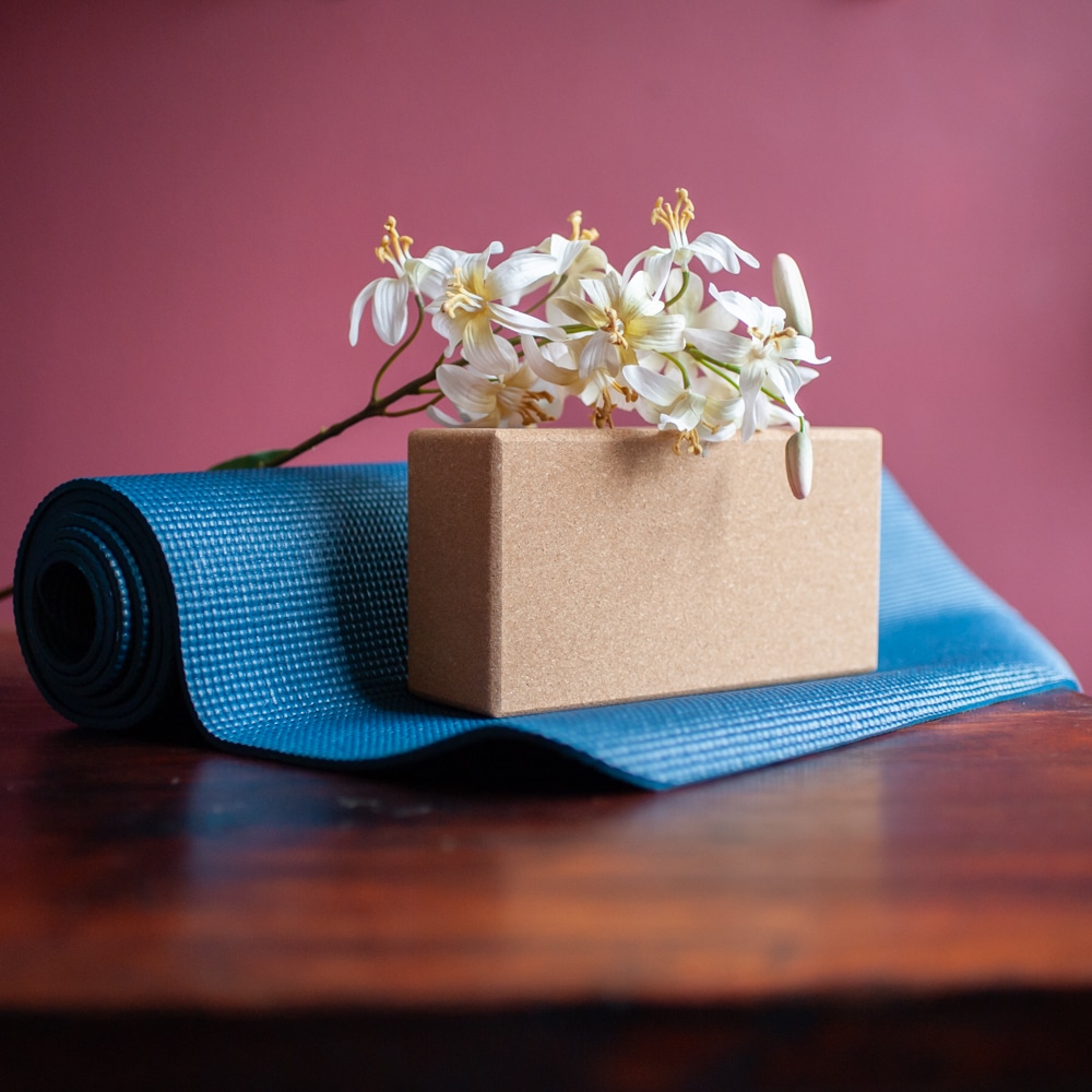 kork yogablock mit blume und aufgerollte blaue Yogamatte auf braunem Holztisch vor roter Wand