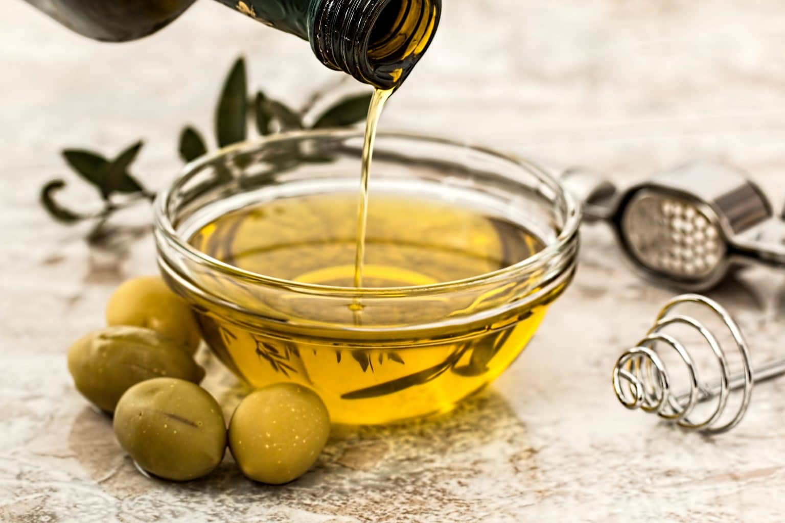 Olivenöl aus Flasche in eine Glasschale mit Oliven und Kräutern und Knoblauchpresse